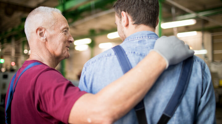Senior man in workwear and gloves touching shoulder of his young subordinate during talk