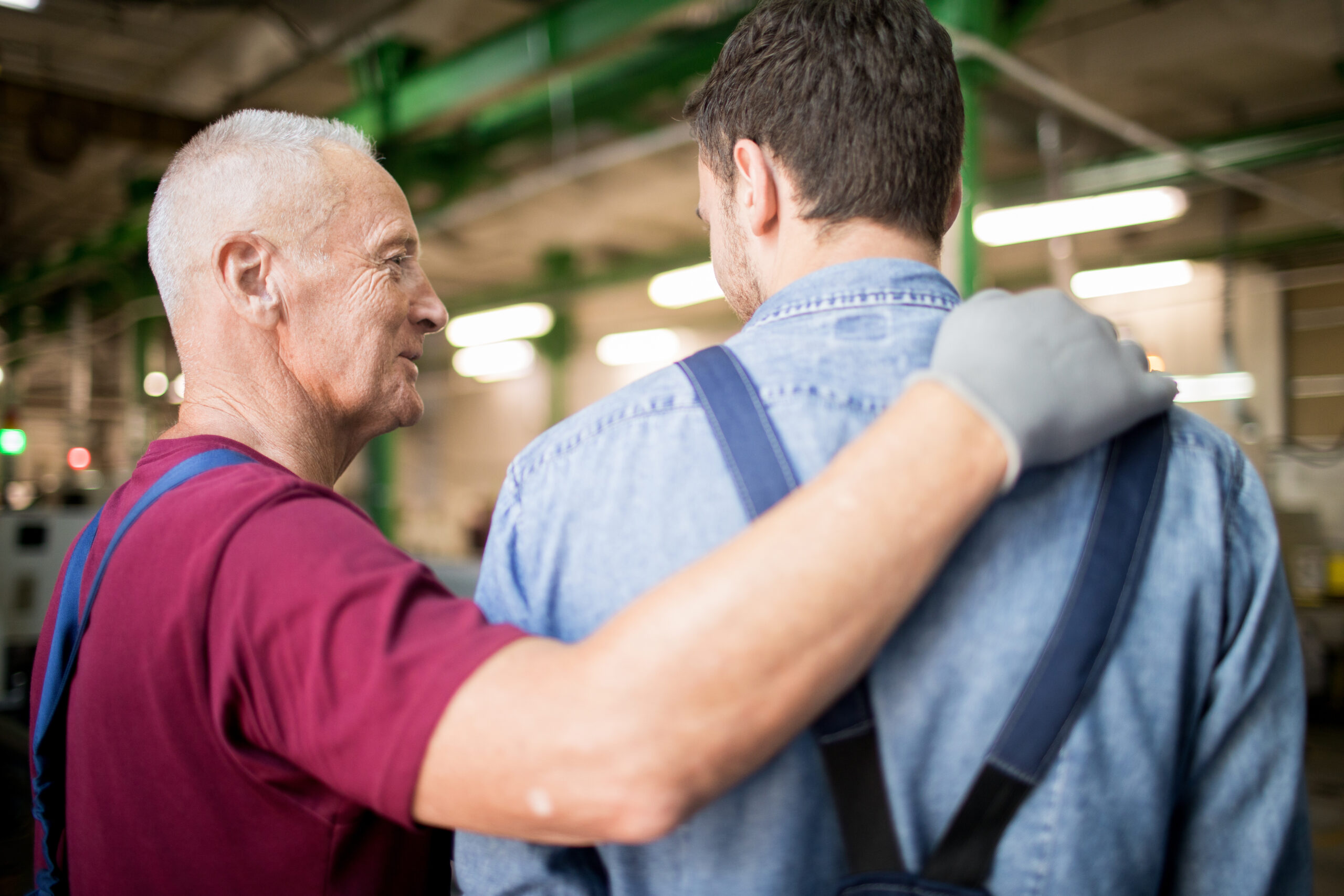 Senior man in workwear and gloves touching shoulder of his young subordinate during talk