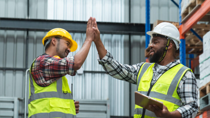 Asian foreman and African engineer in safety hat giving high five and smiling at each other, working at site, listing problem on clipboard to discuss, inspect plan for build a building at warehouse