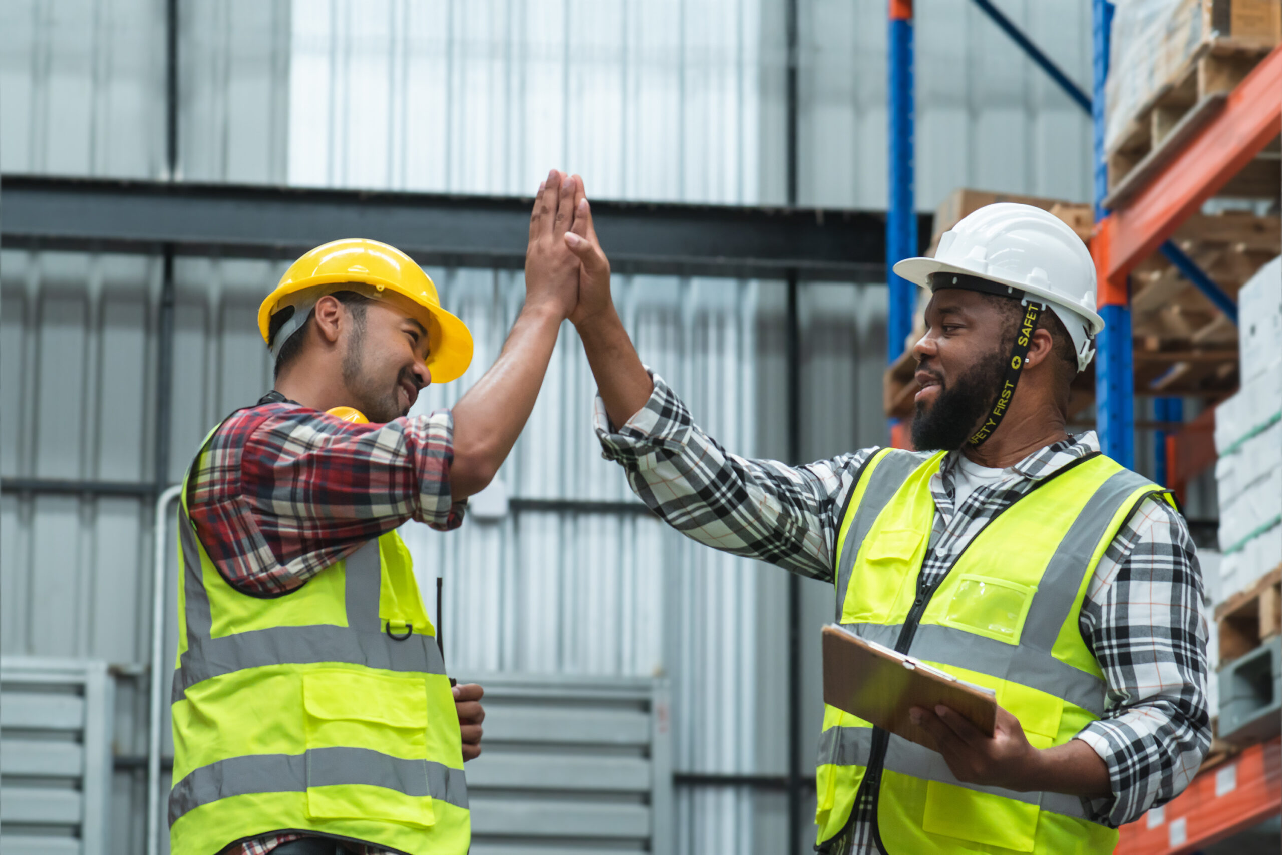 Asian foreman and African engineer in safety hat giving high five and smiling at each other, working at site, listing problem on clipboard to discuss, inspect plan for build a building at warehouse